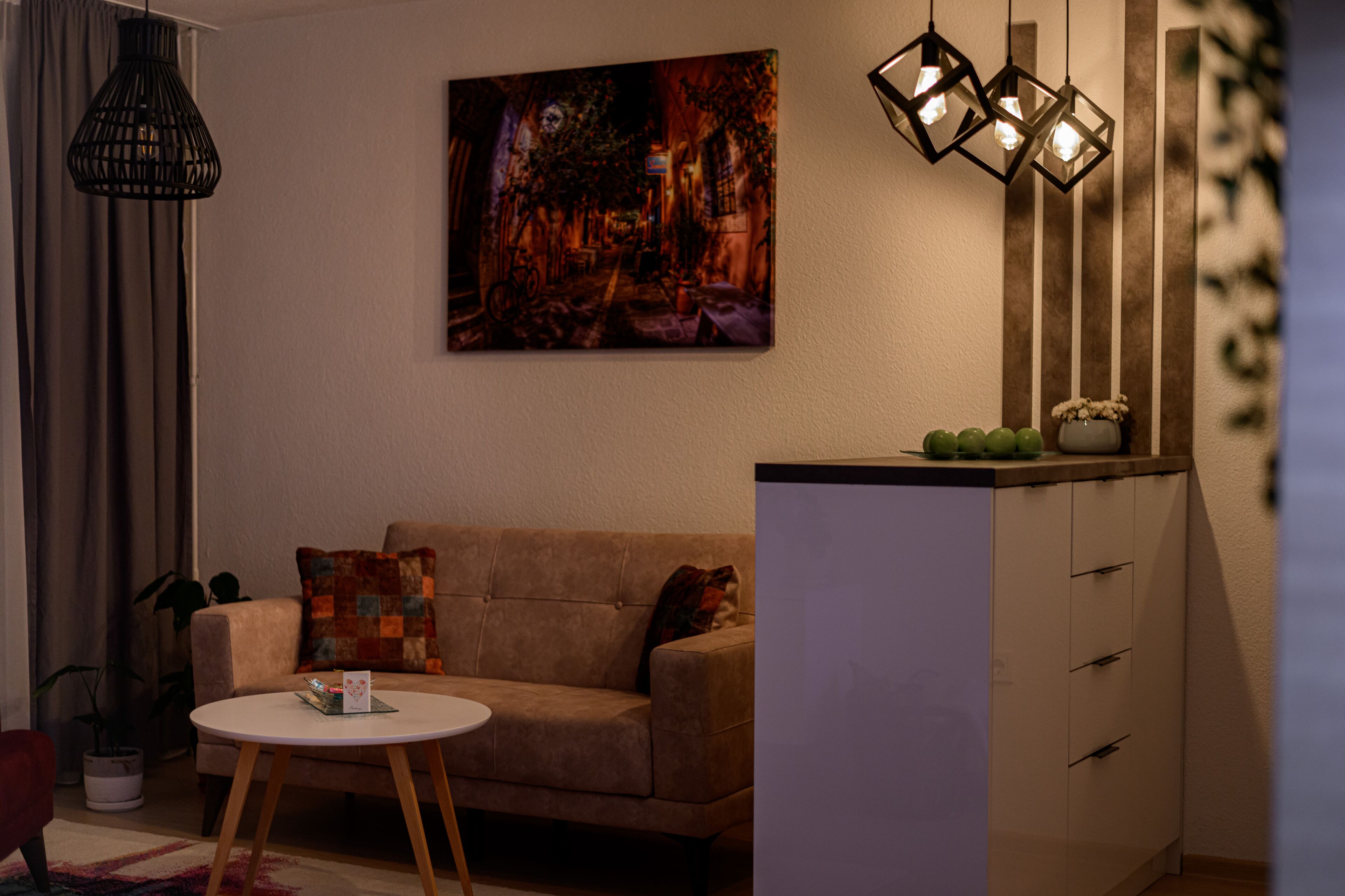 Un salon avec un canapé beige, une table basse ronde et un tableau au mur. On y trouve trois lampes suspendues et un meuble sur lequel est posée une plante.