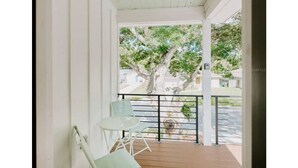 Een overdekt balkon met een tafel, stoelen, een leuning en uitzicht op een boom en gebouwen.