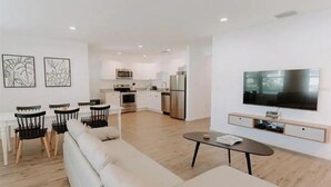 Een moderne woonkamer met een bank, eettafel, stoelen, een flatscreen televisie en een keuken op de achtergrond.