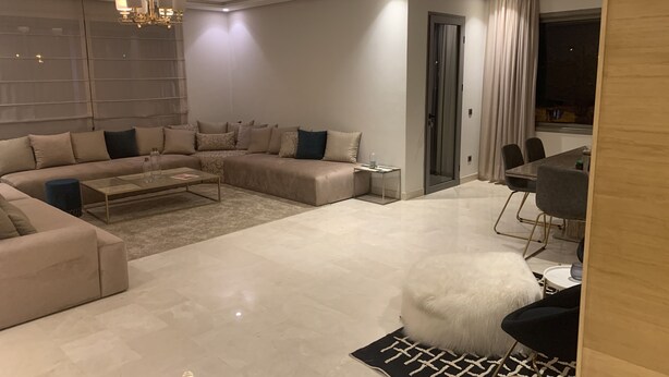 A modern living room with a sectional sofa, a coffee table, a fluffy white pouf, and a dining area with chairs and a table. - Cosy, upscale apartment in California (Casablanca)