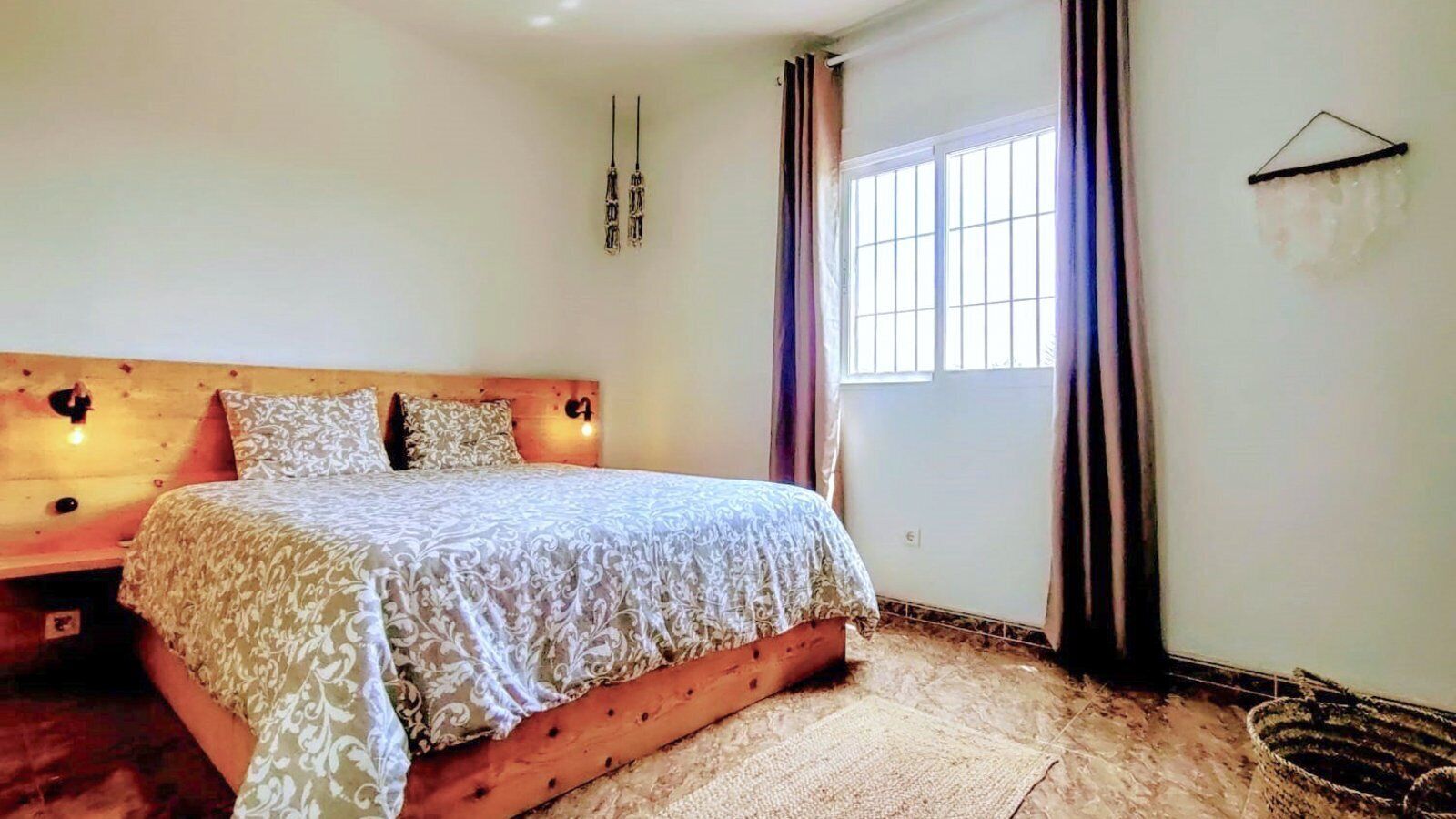 Un dormitorio con estructura de cama de madera, ropa de cama estampada, una ventana con cortinas y un aplique de pared.