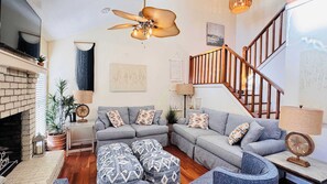 Una sala de estar con chimenea, sofás grises, cojines estampados y una escalera de madera.