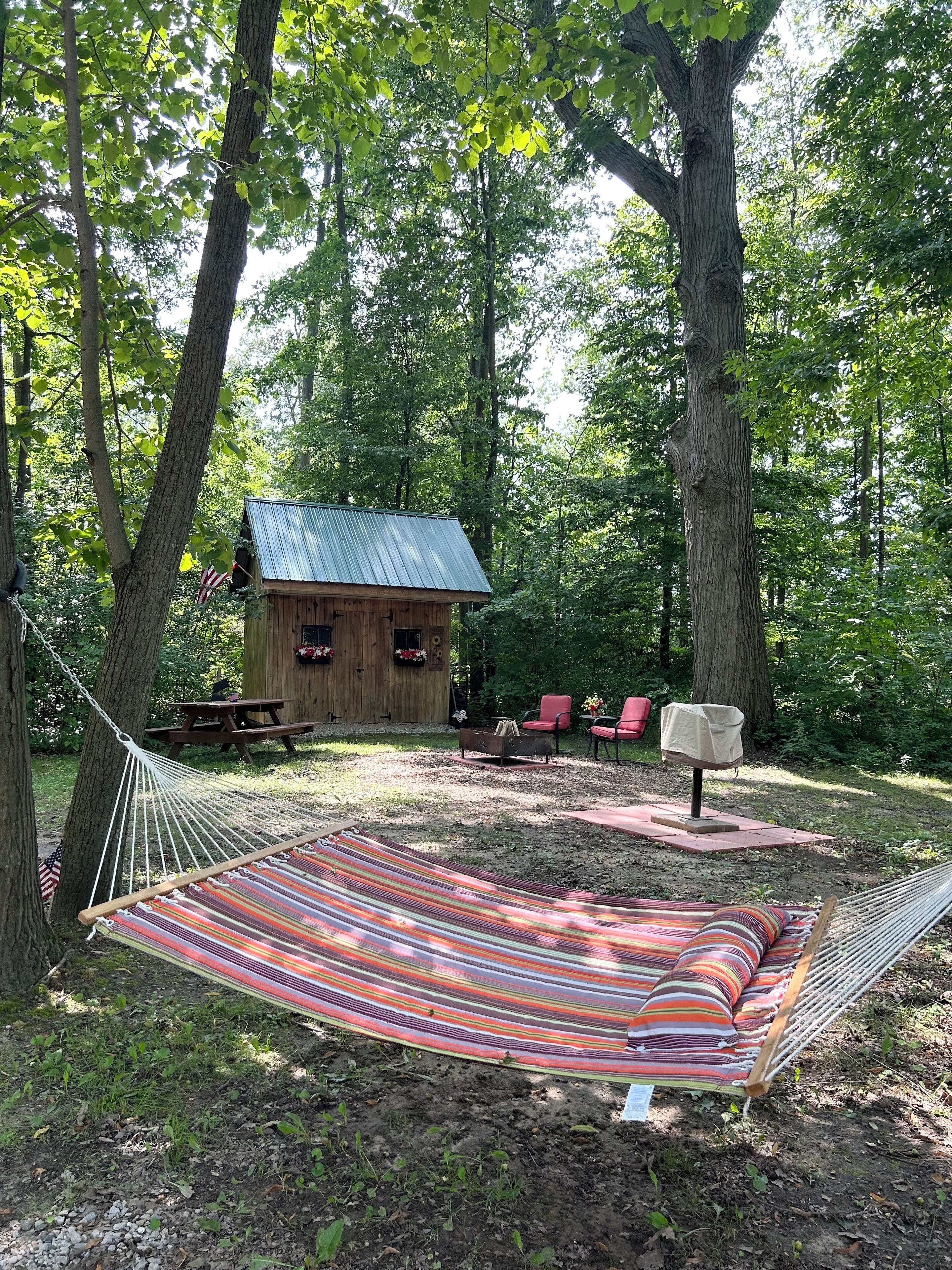 Un hamac dans une zone boisée, avec une petite cabane en bois, une table de pique-nique et des chaises rouges.