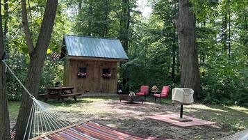 Un hamac dans une zone boisée, avec une petite cabane en bois, une table de pique-nique et des chaises rouges.