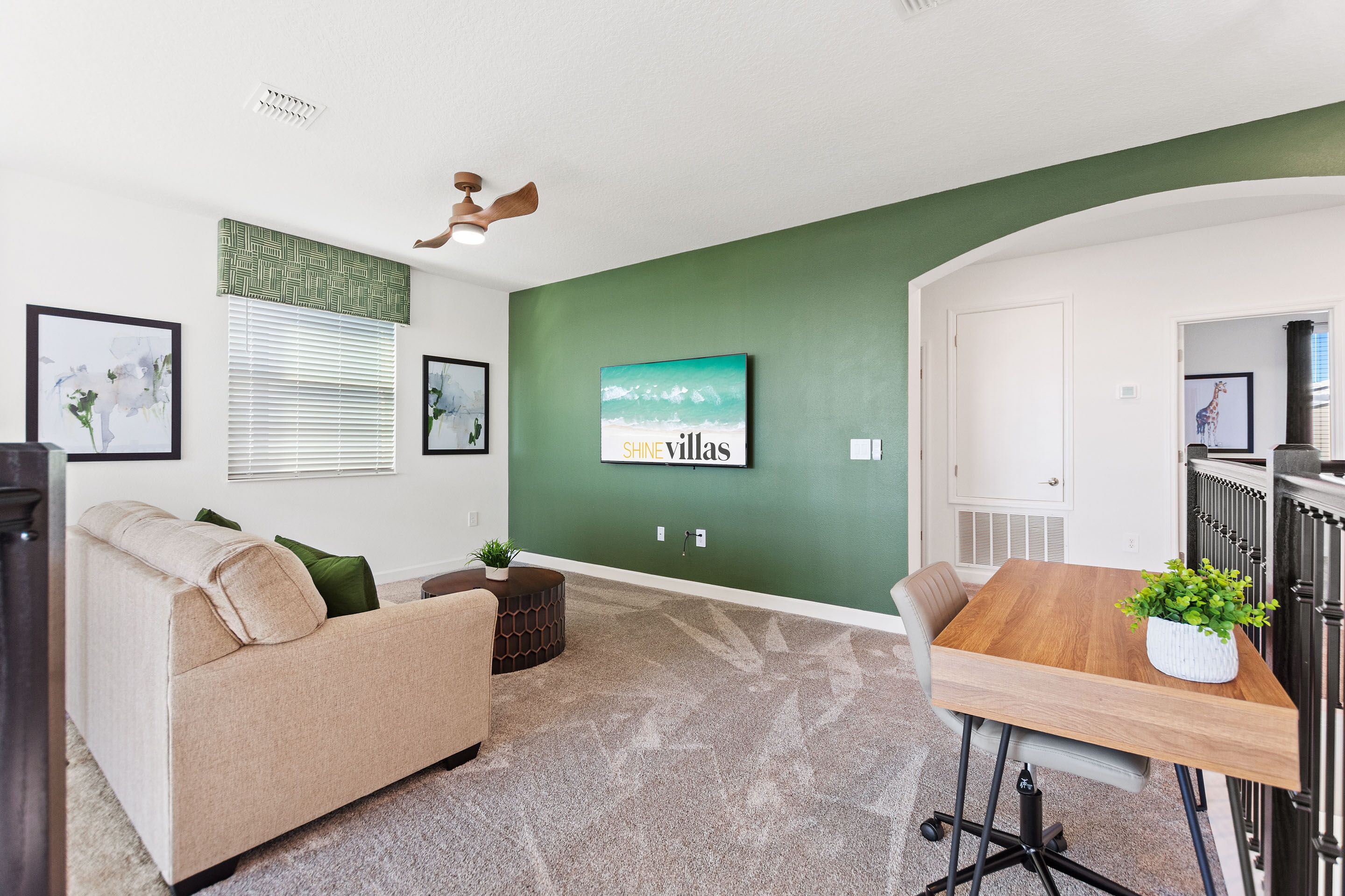A modern living room with a green accent wall, a wooden dining table, a beige sofa, and a ceiling fan.