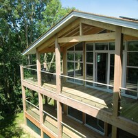 Una terraza de madera con varios niveles y barandillas de vidrio, rodeada de árboles.
