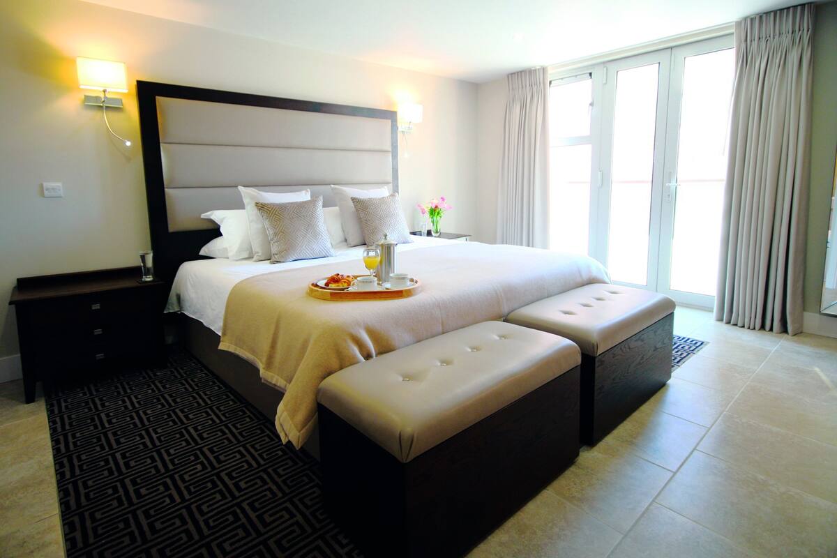 A hotel room with a large bed, a bedside table, a tray with breakfast, and a view of the outdoors.