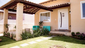 Une maison dotée d’un patio couvert, d’une porte d’entrée avec une plaque indiquant « Résidence », et d’un petit jardin agrémenté de plantes en pot et d’une pelouse.