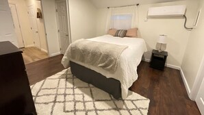 Une chambre à coucher comprenant un lit, une table de chevet, une lampe, une fenêtre avec des rideaux et un appareil de climatisation.