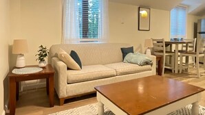 Un salon avec un canapé beige, une table basse en bois, une table d’appoint avec une lampe, et un coin repas comprenant une table et des chaises.