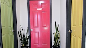 Un pasillo con tres puertas etiquetadas A5, A6 y A7, cada una con un color diferente. Hay dos plantas en macetas a cada lado de la puerta roja central, que tiene un cartel que dice “Solo personal”. Dos felpudos en el suelo dicen “Nuestros huéspedes”.