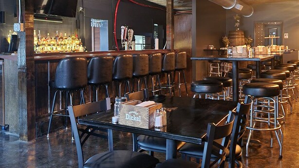 Un restaurante con una decoración rústica, que incluye vigas de madera a la vista, una zona de bar con taburetes y una larga mesa de comedor.
