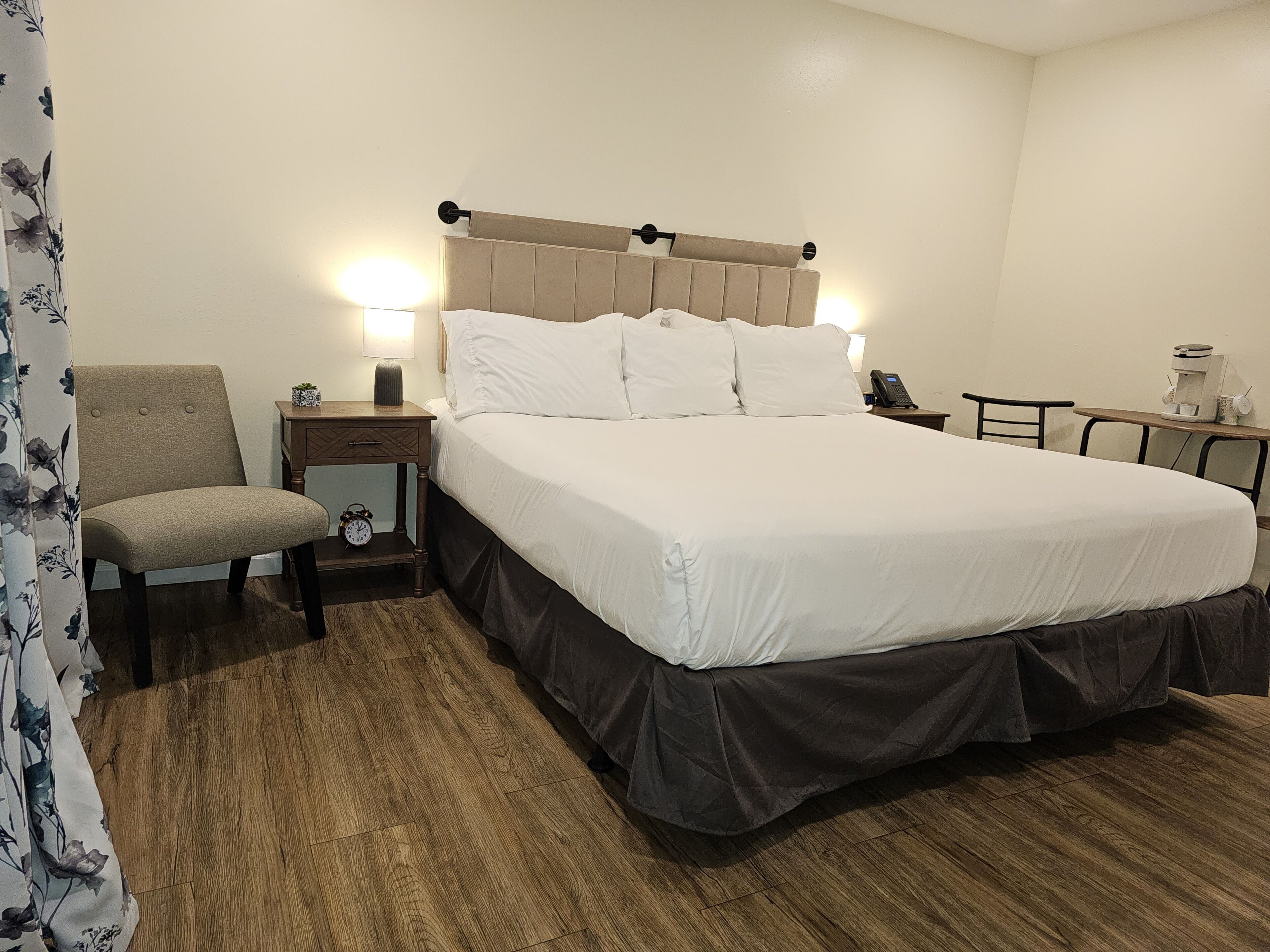 Habitación de hotel con cama, una silla, mesita de noche con lámpara, cafetera y un reloj.