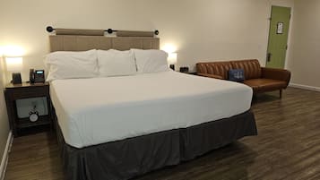 A hotel room with a large bed, a nightstand with a clock, a lamp, and a green door.
