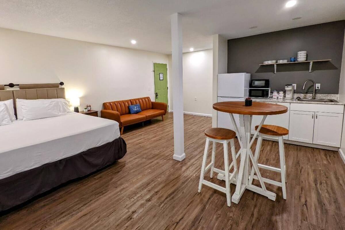 Habitación de hotel con cama, sofá, mesa de comedor con taburetes, kitchenette y una puerta verde.