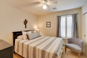 Ein Schlafzimmer mit einem Bett, einem Sessel, einem Deckenventilator, einem Fenster mit Vorhängen und einer Kakteendeko an der Wand.