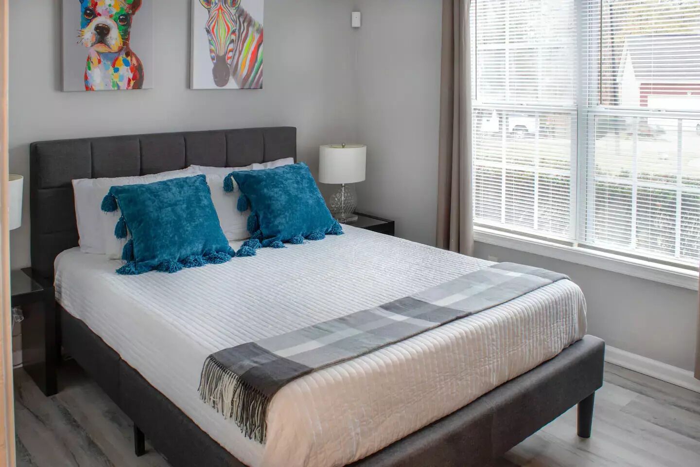 Un dormitorio moderno con una cama grande, dos almohadas azules, un edredón blanco y una manta gris. Hay dos obras de arte en la pared sobre la cama, una ventana con persianas y una mesita de noche con una lámpara.