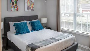 A modern bedroom with a large bed, two blue pillows, a white duvet, and a grey throw. There are two wall art pieces above the bed, a window with blinds, and a nightstand with a lamp. - Private basement w/firepit ideal for longer stays (Suwanee)