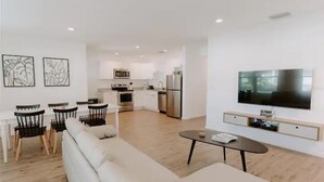 Een moderne woonkamer met een bank, een salontafel en een aan de muur bevestigde televisie. In de achtergrond is een keuken te zien met een magnetron, oven en een koelkast.