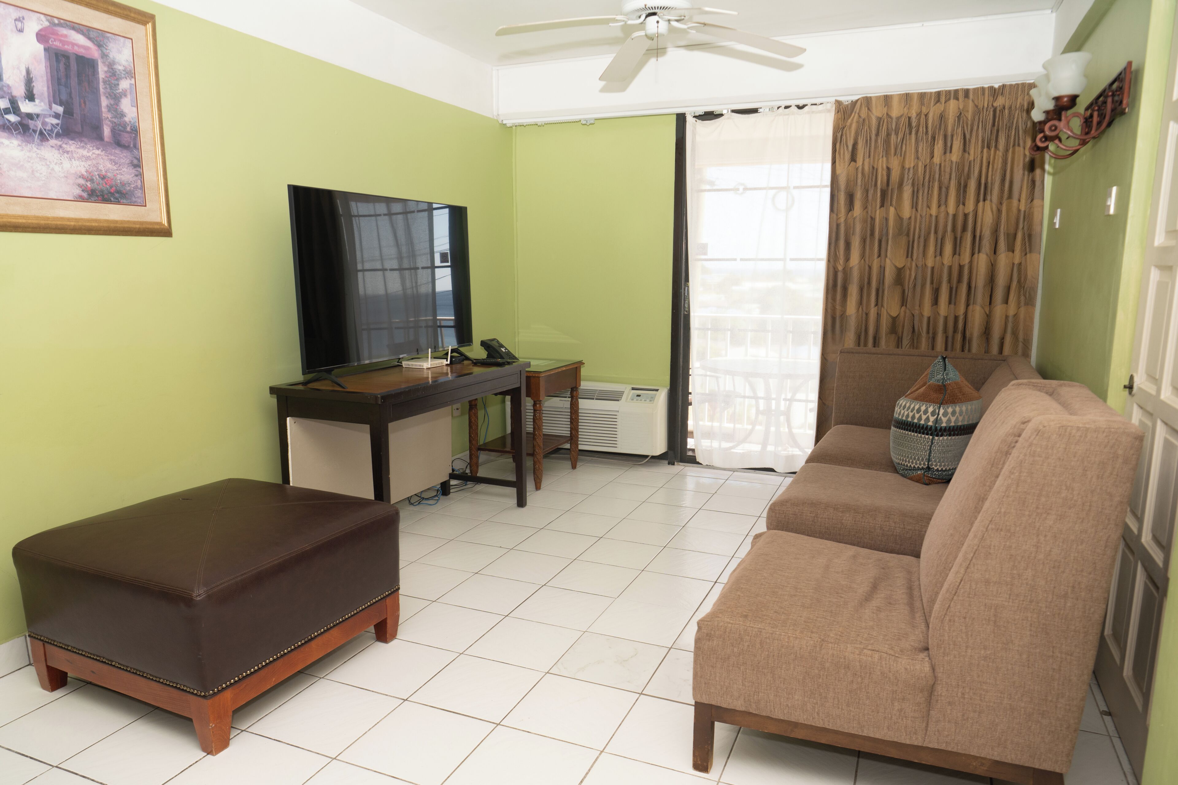 A living room with a sofa, ottoman, TV, desk, and a painting on the wall.