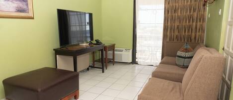 A living room with a sofa, ottoman, TV, desk, and a painting on the wall.