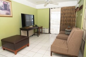 A living room with a sofa, ottoman, TV, desk, and a painting on the wall.