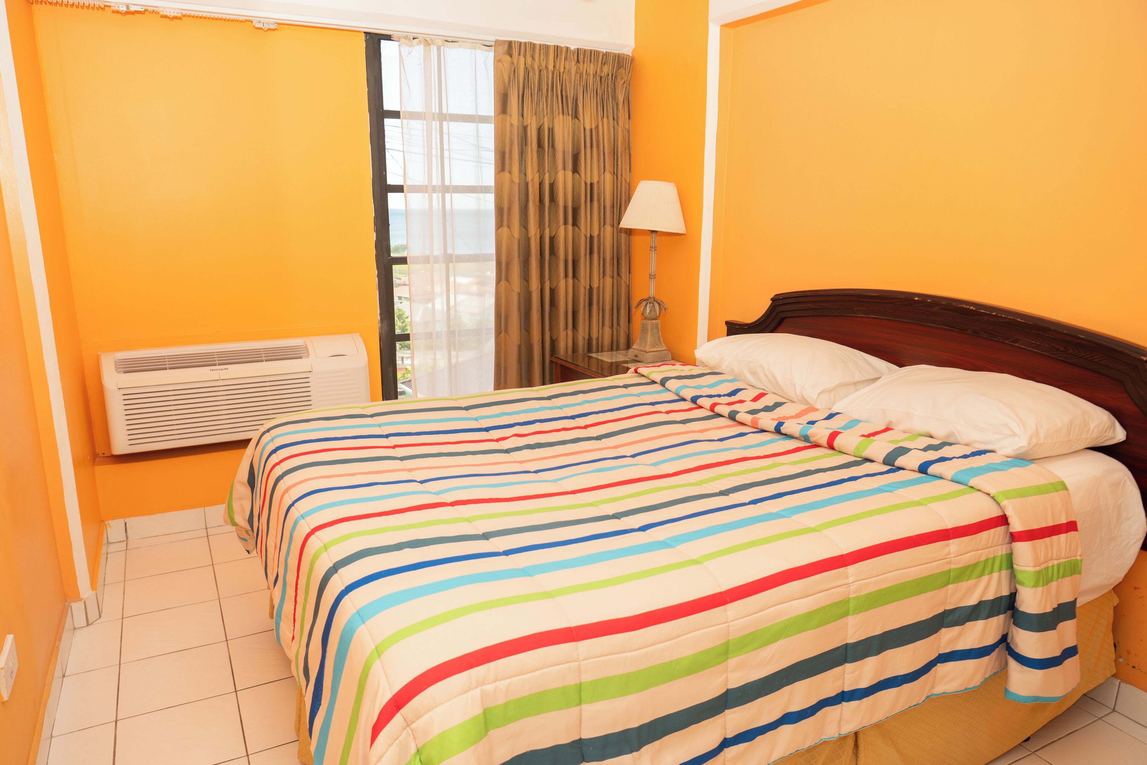A hotel room with a bed, a window with curtains, a bedside lamp, and an air conditioning unit.