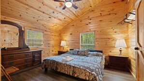 Dormitorio de cabaña de madera con cama, dos mesitas de noche, cómoda, ventilador de techo y dos ventanas con persianas.