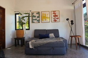 A modern bedroom with a grey sofa bed, a large potted plant, abstract wall art, and a tripod lamp.