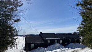 A two-story house with a dark roof and multiple windows, surrounded by snow-covered ground and trees.