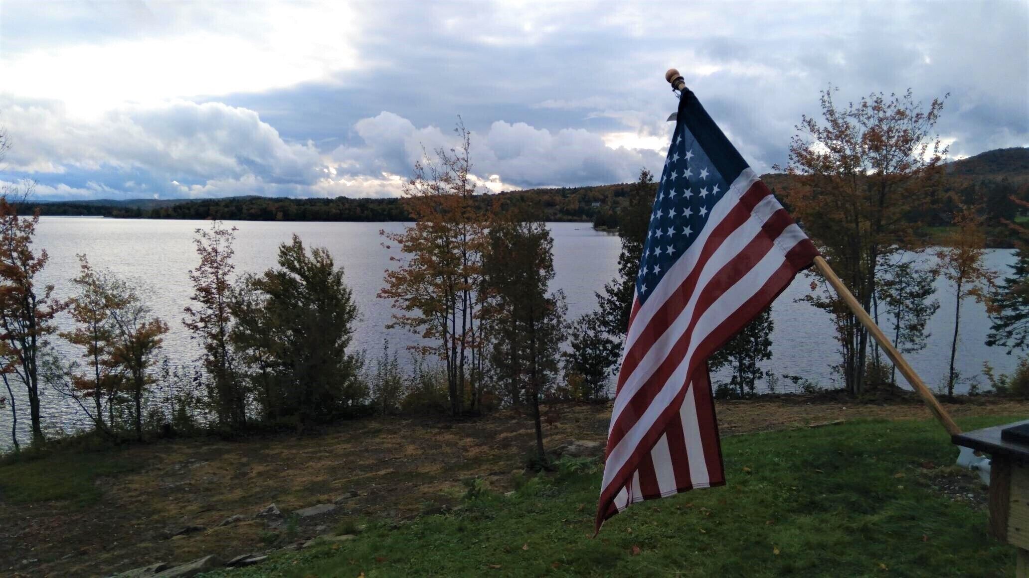 Una bandera estadounidense en un asta, cerca de un lago con árboles y cielo nublado.