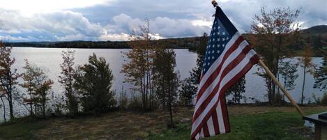 Una bandera estadounidense en un asta, cerca de un lago con árboles y cielo nublado.
