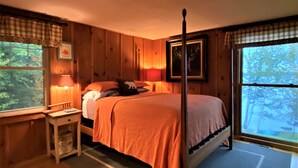 A cozy bedroom with a four-poster bed, a bedside table with a lamp, and a window with a view of trees.
