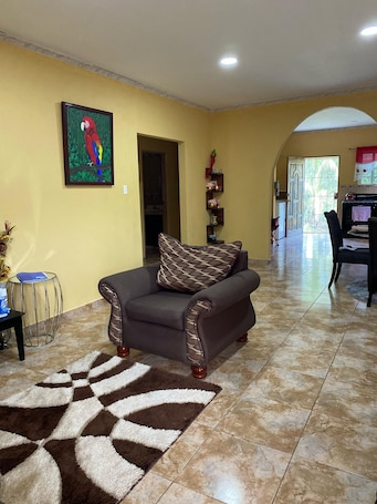 A living room with a sofa, a patterned rug, a painting, and a kitchen area visible through an archway.