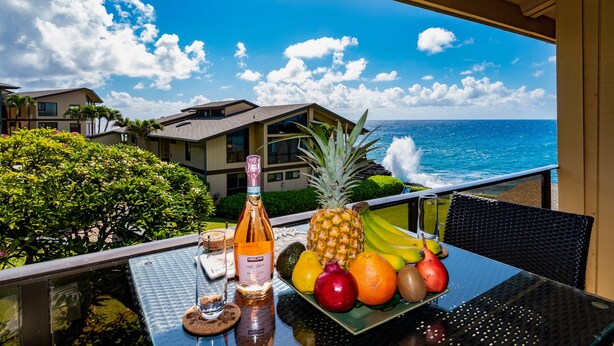 Ein Balkon mit Tisch, gedeckt mit Früchten und einer Flasche, mit Blick auf die Küste, Gebäude und das Meer.