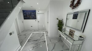 A modern hallway with a marble floor, a staircase, a console table with a mirror, and a cityscape painting on the wall. - Cheerful 4 Bedroom Luxury house with free parking (Halesowen)