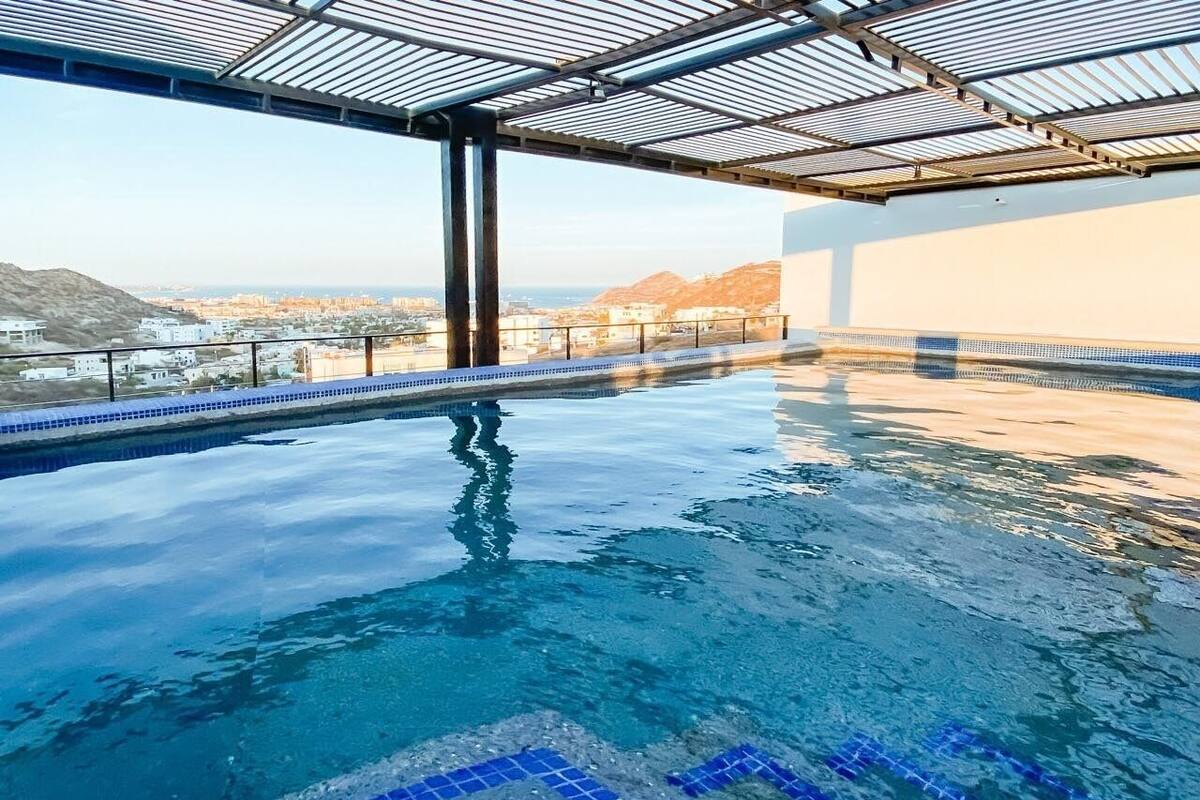 An indoor infinity pool with clear blue water, surrounded by a glass ceiling and a view of distant buildings and hills.