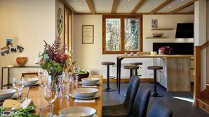 Un comedor con una mesa de madera dispuesta para una comida, taburetes de bar y una cocina al fondo.