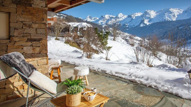 Un edificio de piedra con una terraza de madera, una silla, una mesita con una planta y un bol de aperitivos, y vistas a montañas cubiertas de nieve.