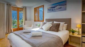 Un dormitorio con una cama grande, una silla, una mesita de noche con lámpara y una ventana con vistas a las montañas.