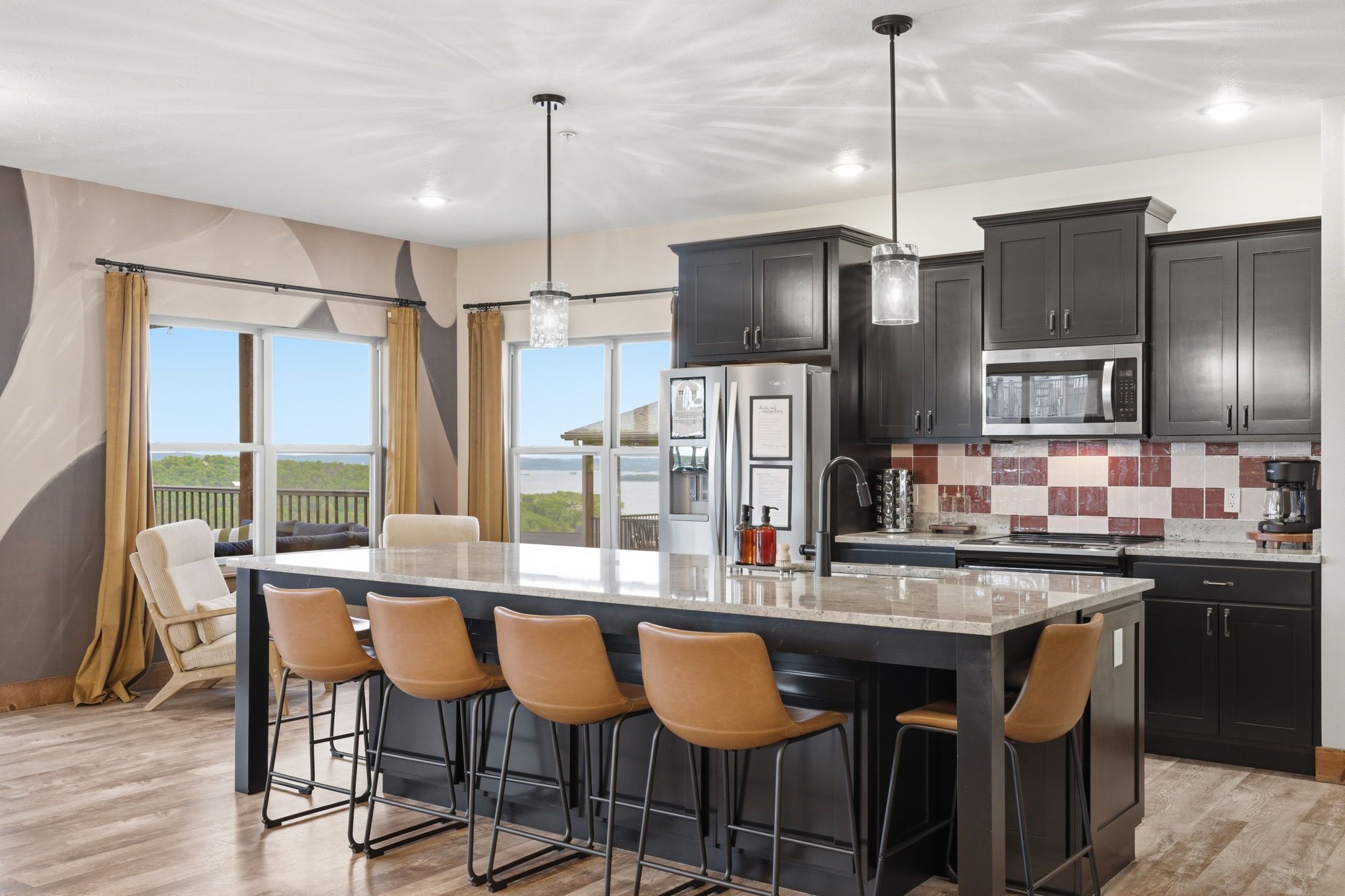 A modern kitchen with a large island, bar stools, dark cabinets, and a view through the window.