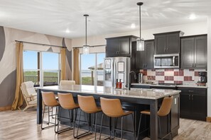 A modern kitchen with a large island, bar stools, dark cabinets, and a view through the window.