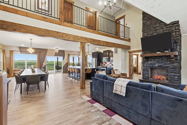 A spacious living area with a high ceiling, a stone fireplace, a dining area with a long table, and a view of the outdoors.