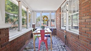 Une véranda avec un mur en briques, de grandes fenêtres, une table avec des chaises et une décoration en forme de couronne.