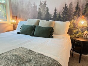 A bed with white bedding and green and white pillows, a grey throw blanket, and a forest-themed wall mural. There are two bedside lamps with woven shades. - Cl113 Moonstone Acre (Columbia Falls)