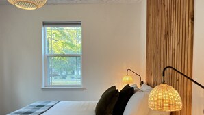 A bedroom with a large bed, a window with a view of trees, a wooden wall, and a lamp. - Cl113 Moonstone Acre (Columbia Falls)