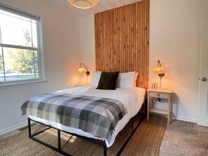 A bedroom with a bed, two bedside lamps, a window, and a wooden wall panel. - Cl113 Moonstone Acre (Columbia Falls)