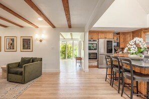 A spacious living room with a green sofa, wooden beams on the ceiling, and a kitchen with wooden cabinets and stainless steel appliances.