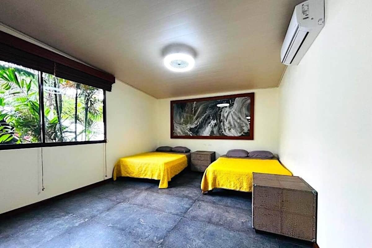 A room with two beds, a large window with a view of greenery, a ceiling fan, and a painting on the wall.