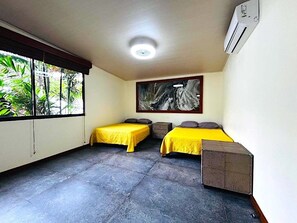 A room with two beds, a large window with a view of greenery, a ceiling fan, and a painting on the wall. - Arenal Backpackers Resort (La Fortuna)